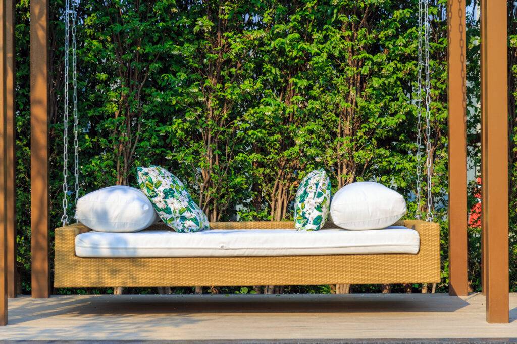 An outdoor resin wicker porch swing in the garden with nature ba bed swing in a lush garden on a high anchor point with pristine white bed swing covers